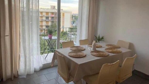 A dining area with a table set for six, large windows with sheer curtains, and a view of a residential area. - T3 Marseille 10mn drive from Velodrome & beach (Marseille)