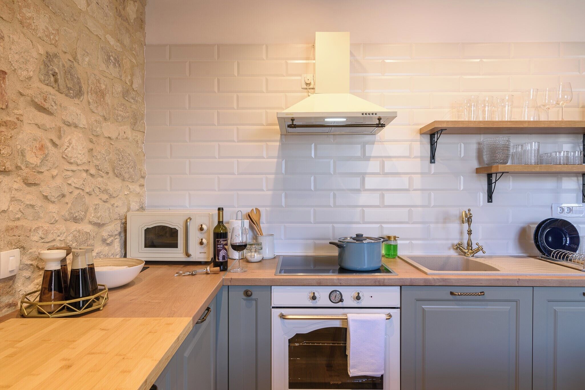 A kitchen with a stone wall, white tiled backsplash, a stove, oven, and a sink.
