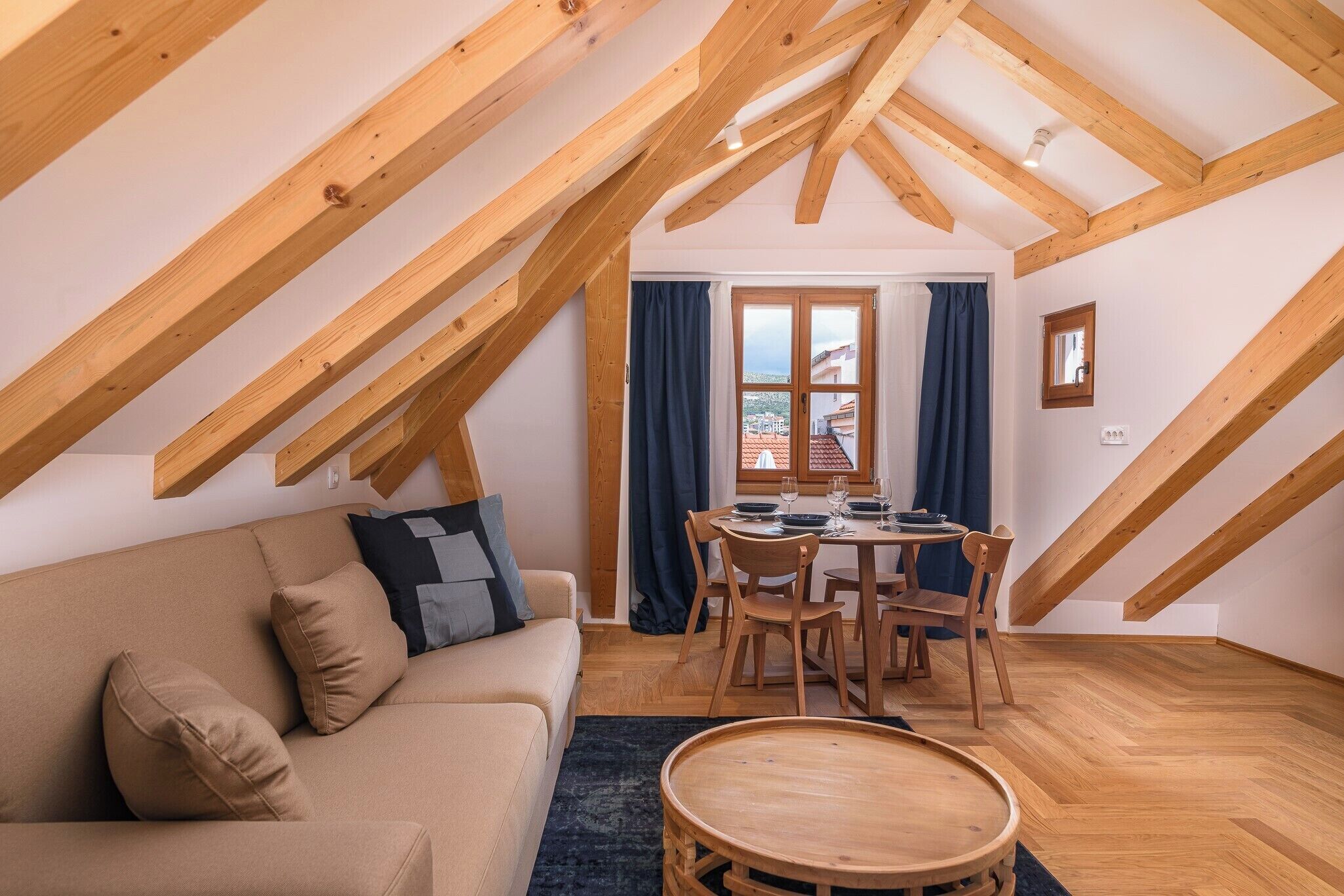A cozy loft-style room with a wooden ceiling, a sofa, a dining table, and a view through a window.