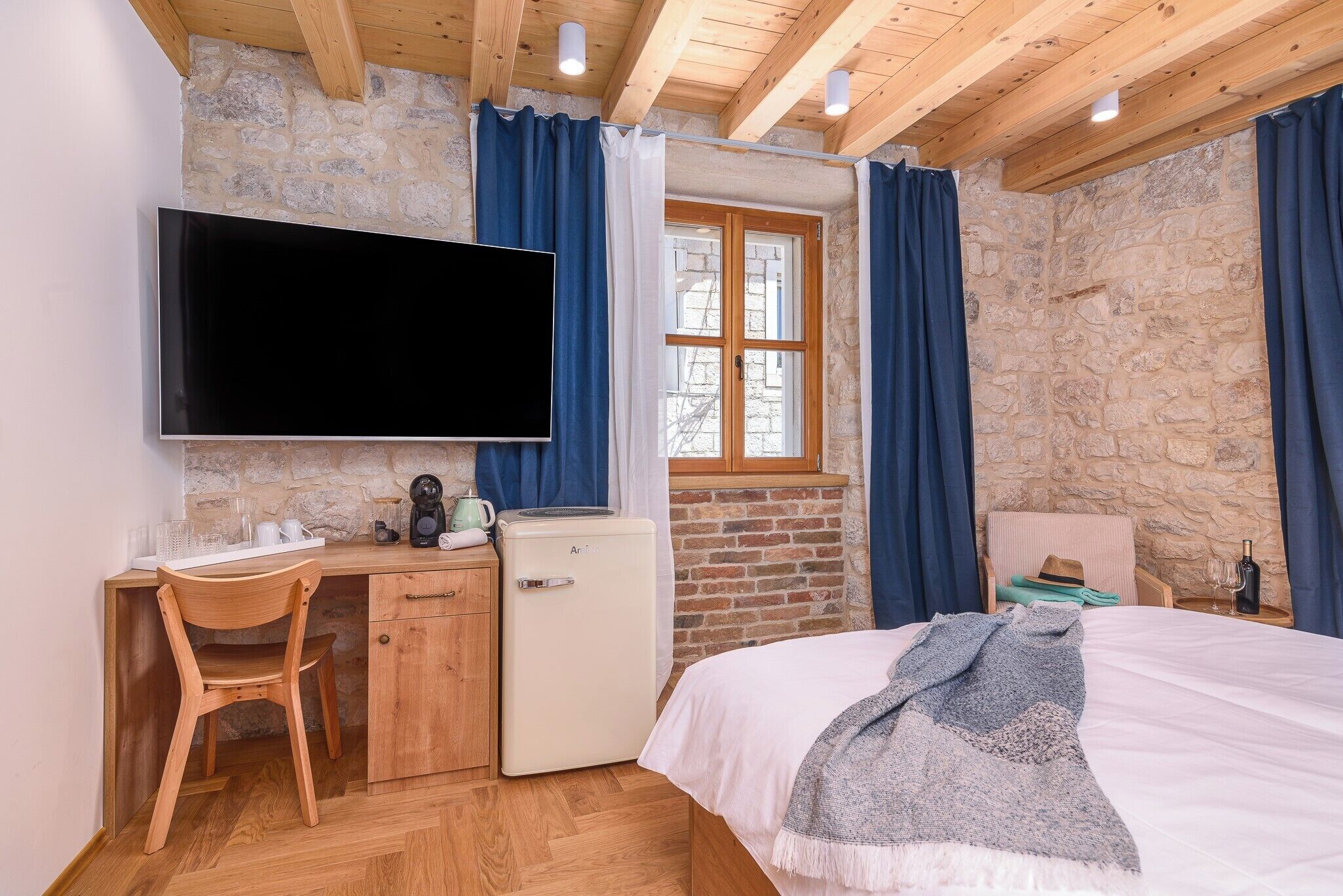 Un dormitorio con cama, escritorio y silla, televisor, refrigerador pequeño y ventana con cortinas.