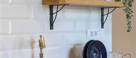 A kitchen with a wooden countertop, a sink with a brass faucet, and two shelves above holding glassware and a plant.