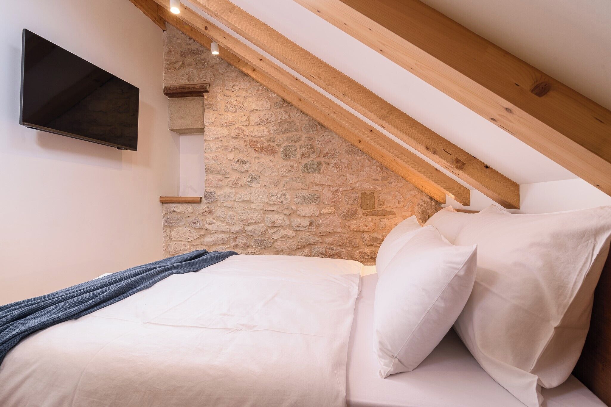 Quarto com parede de pedra, vigas de madeira, televisão de tela plana e cama com roupa de cama branca.