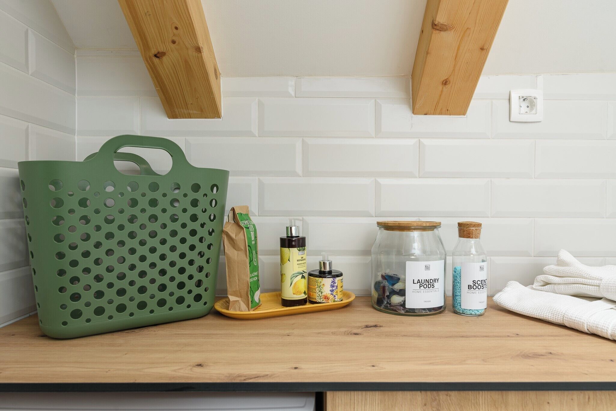 A kitchen counter with a green basket, a wooden spoon, a tray with bottles, and a jar.