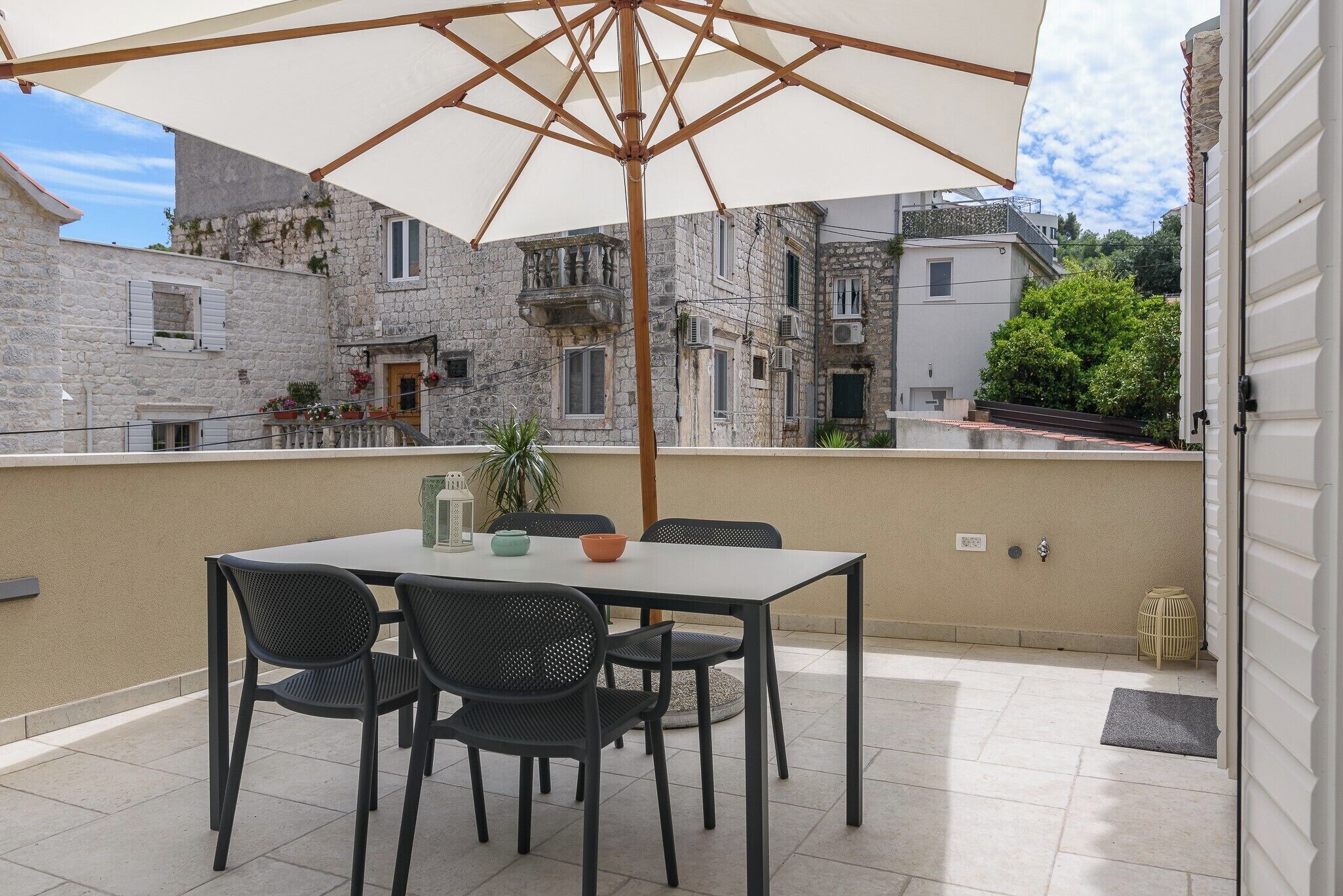 A terrace with a table and chairs under a large umbrella, overlooking a historic stone building with balconies and greenery.