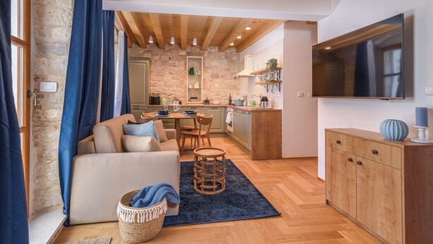 A cozy living room with a sofa, a coffee table, and a TV. There is a kitchen area with a dining table and chairs. - Oleander Center Suites (Trogir)
