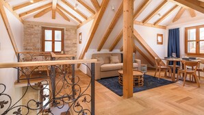 A spacious loft with exposed wooden beams, a stone wall, a staircase with wrought iron railings, a sofa, a dining table with chairs, and a large window with curtains. - Oleander Center Suites (Trogir)