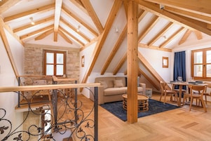 Un loft espacioso con vigas de madera a la vista, una pared de piedra, una escalera con barandillas de hierro forjado, un sofá, una mesa de comedor con sillas y un amplio ventanal con cortinas.