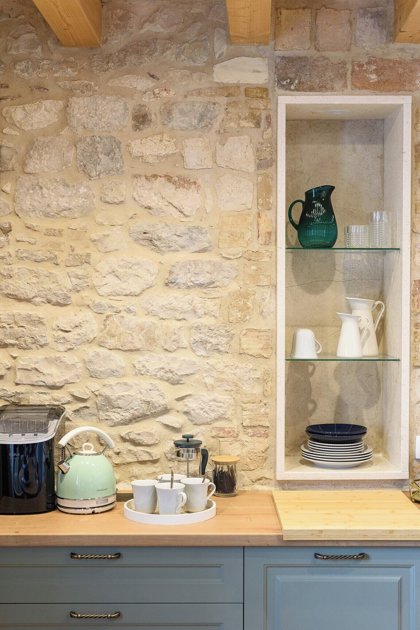 A kitchen with a stone wall, a wooden countertop, a tea kettle, a coffee maker, and a glass cabinet with dishes.