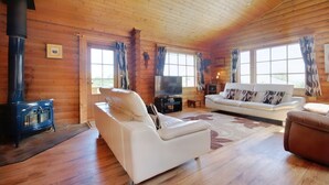 Interior de una cabaña de madera con un sofá blanco, un televisor y un estufa de leña.