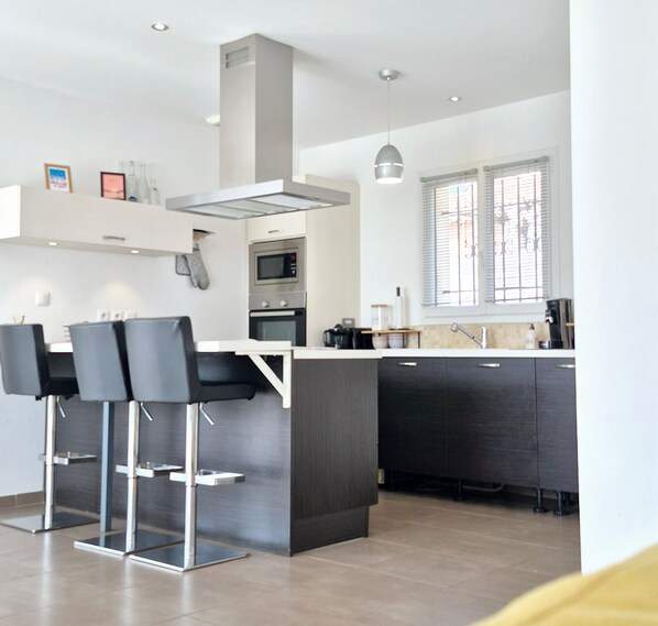 A modern kitchen with a central island, bar stools, a built-in oven, and a window with blinds. - Villa Avec Piscine au Coeur D'une Station Balnéaire (La Seyne-sur-Mer)