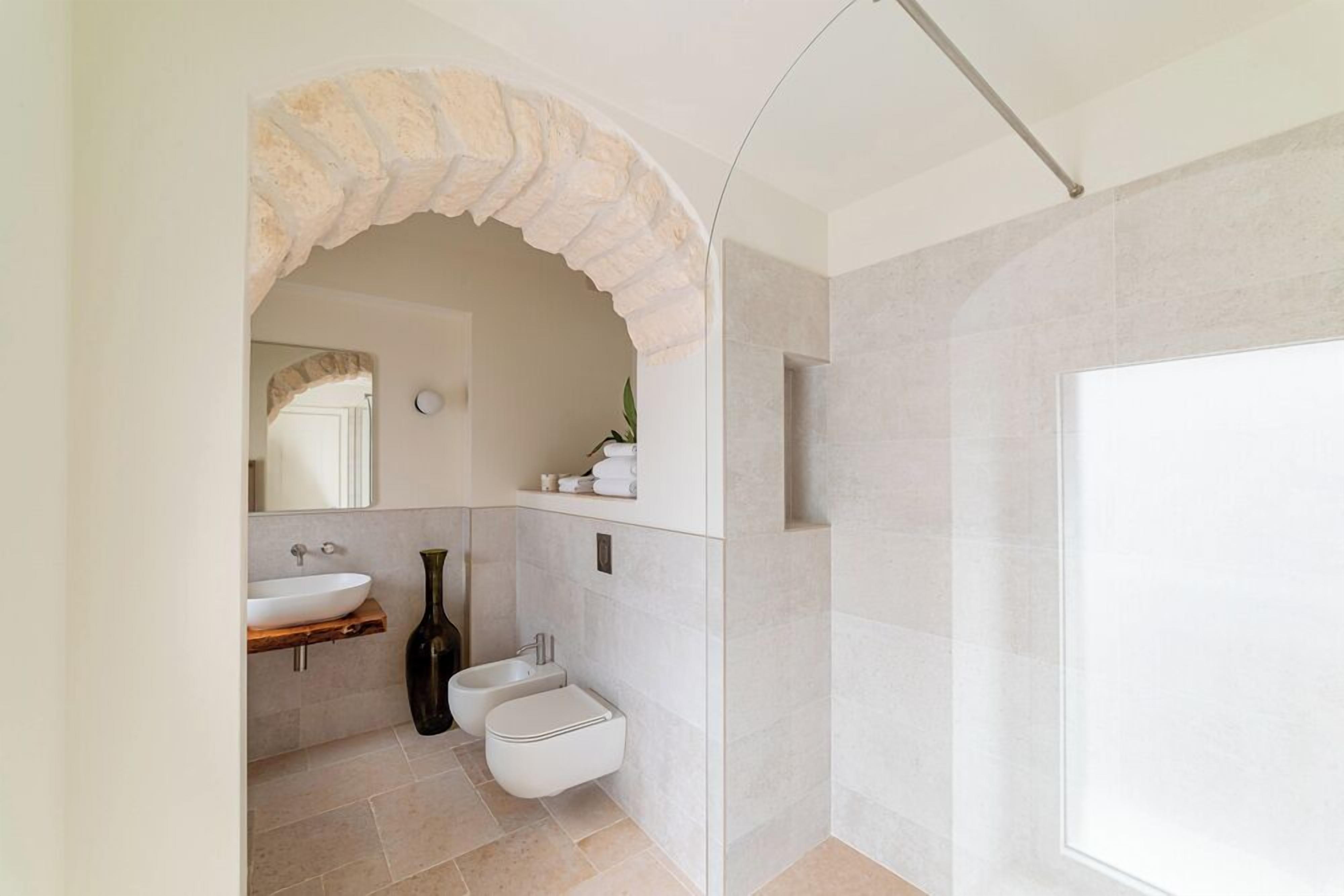 Un baño moderno con un arco de piedra, un lavamanos blanco, un bidé y una zona de ducha.