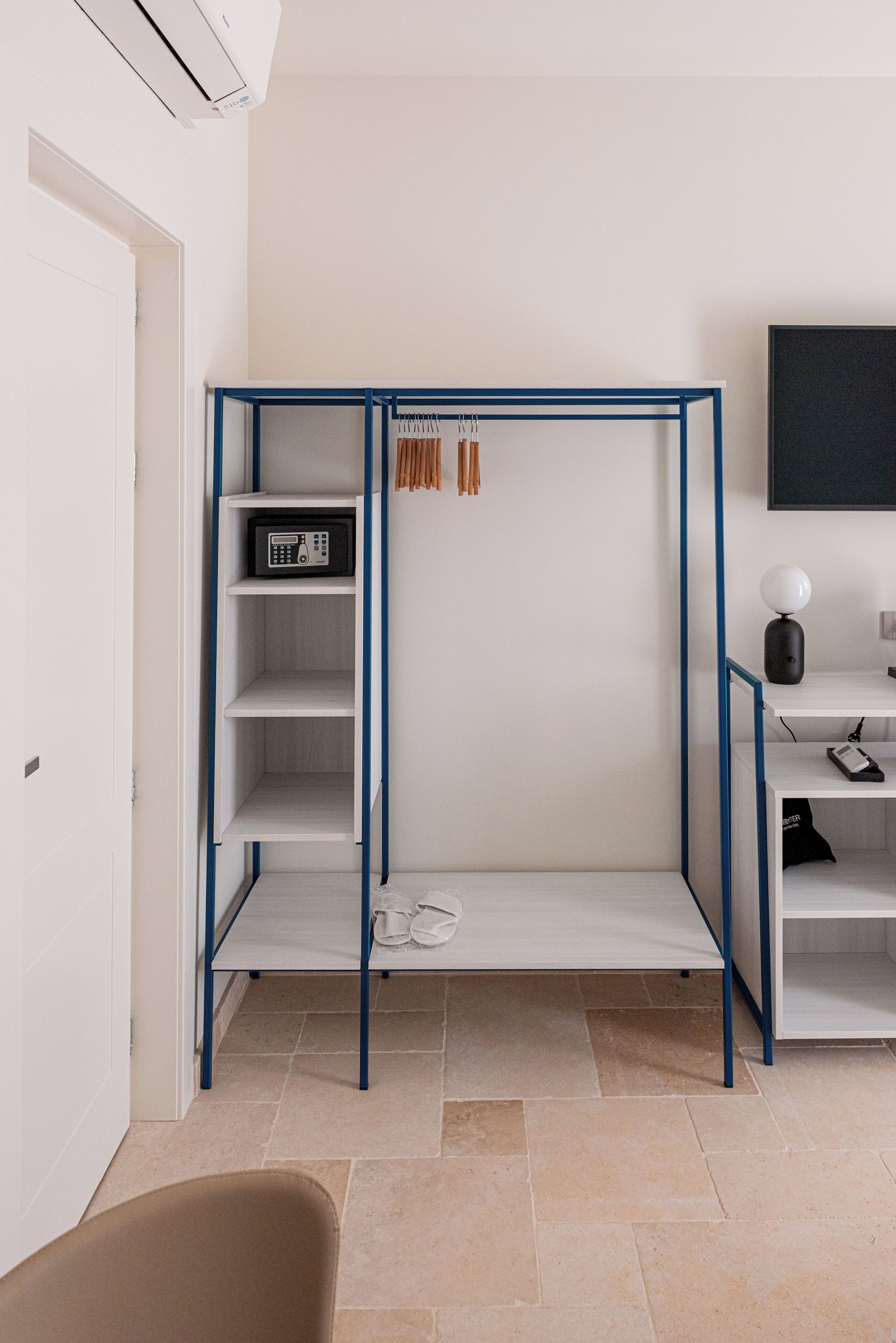Une pièce avec une armoire blanche aux montants bleus, une petite étagère avec une radio, une paire de chaussons et un bureau avec une lampe et un ordinateur.