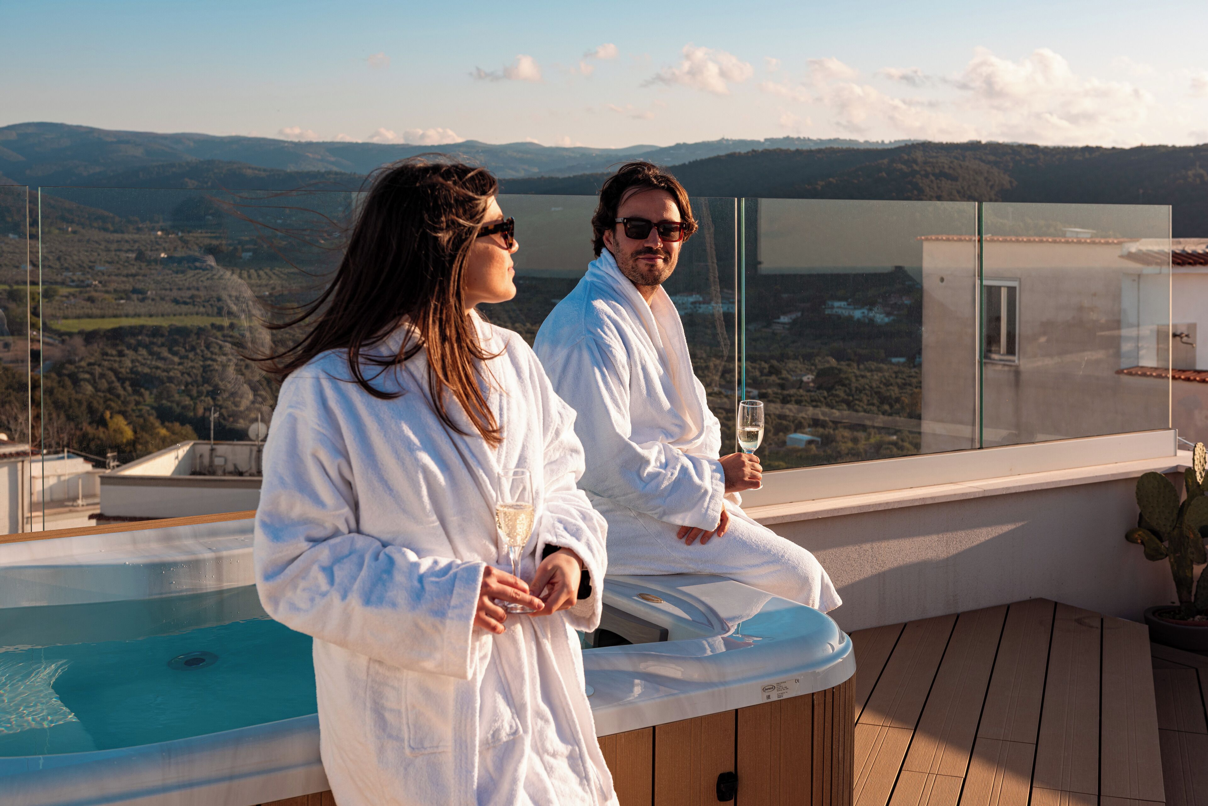 A couple in white robes on a balcony with a hot tub and a view of the countryside.