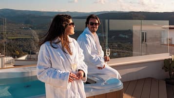 A couple in white robes on a balcony with a hot tub and a view of the countryside.