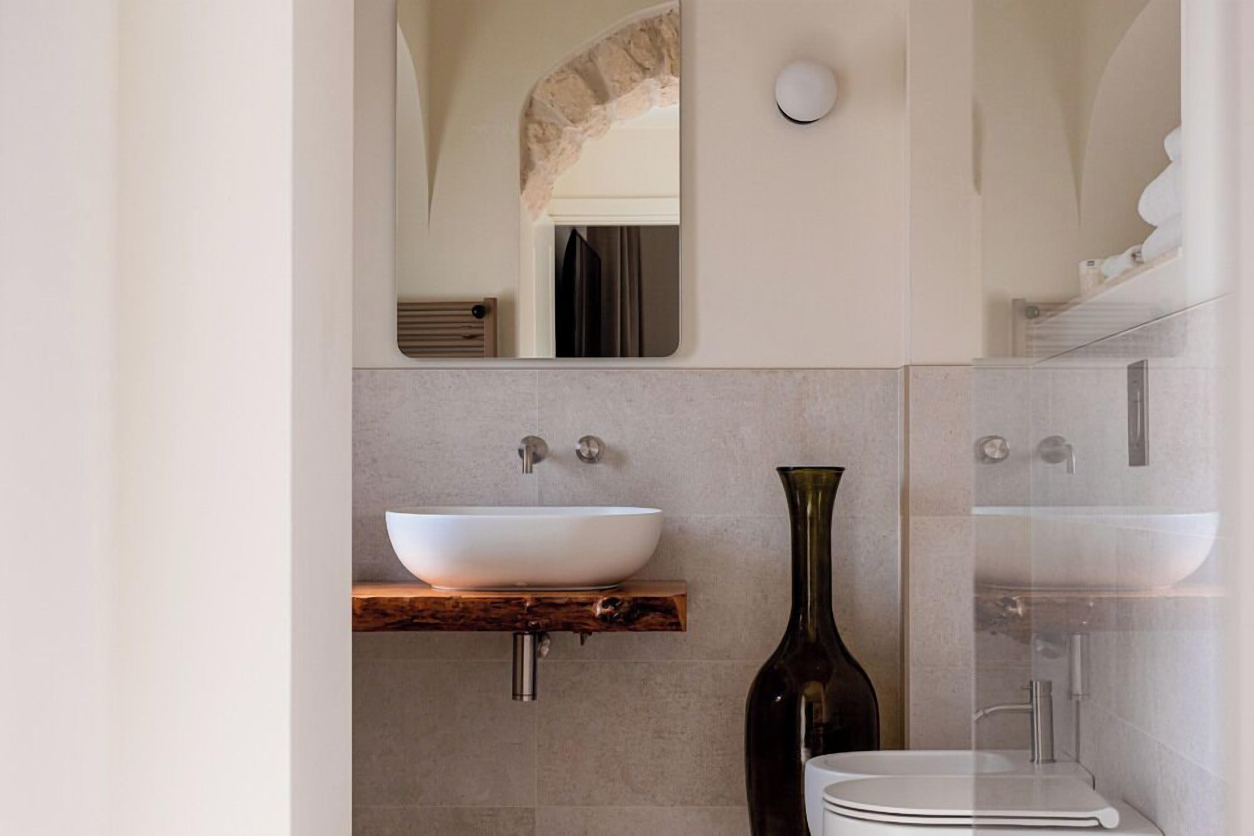 Un baño moderno con un lavabo blanco tipo recipiente, un estante de madera, un jarrón oscuro y una ducha con cerramiento de vidrio.