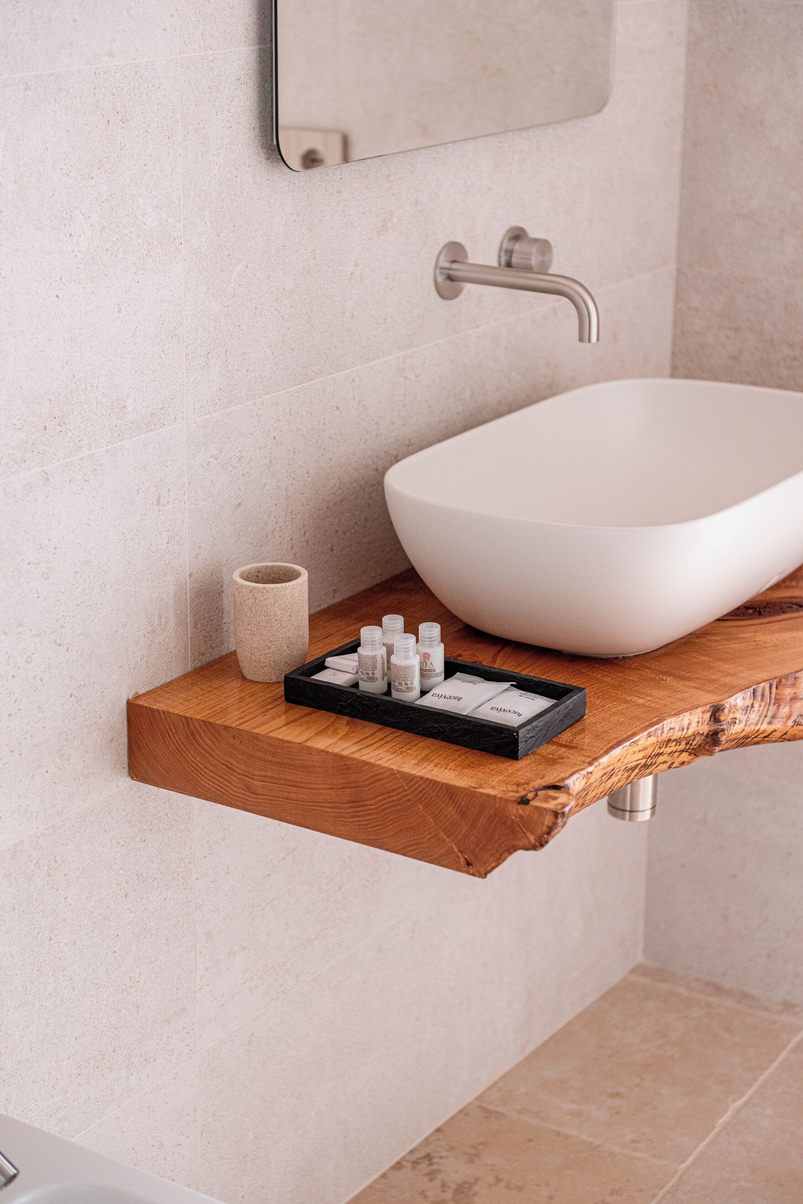 A bathroom with a white vessel sink, a wooden shelf with toiletries, and a textured wall.
