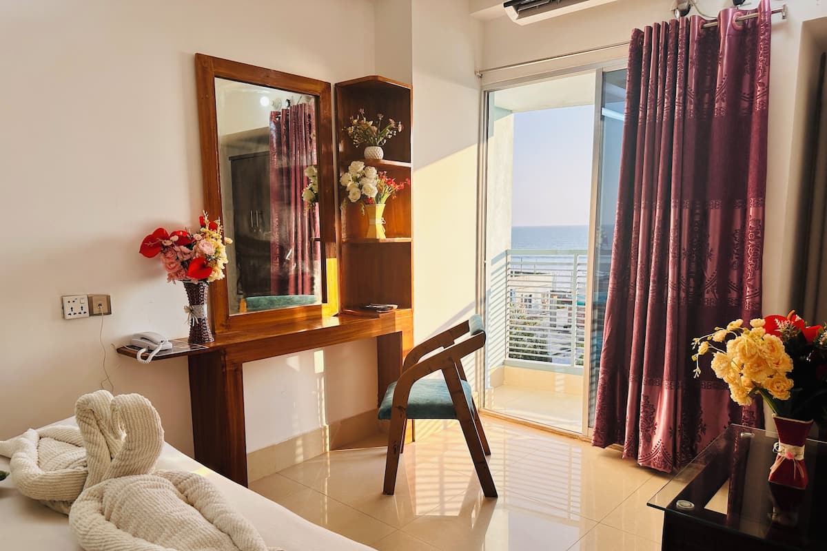 Habitación de hotel con cama, escritorio con espejo, florero con flores y vista al mar a través de una puerta corredera.