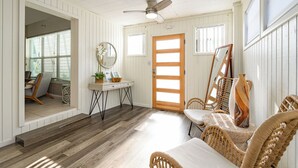 Una habitación con piso de madera, paredes blancas, un ventilador de techo, una puerta de madera, una mesa con un florero y sillas de mimbre.