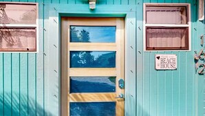 Una casa de playa con puerta de madera, un cartel que dice 'Welcome to the Beach House' y una ventana con vista a árboles.