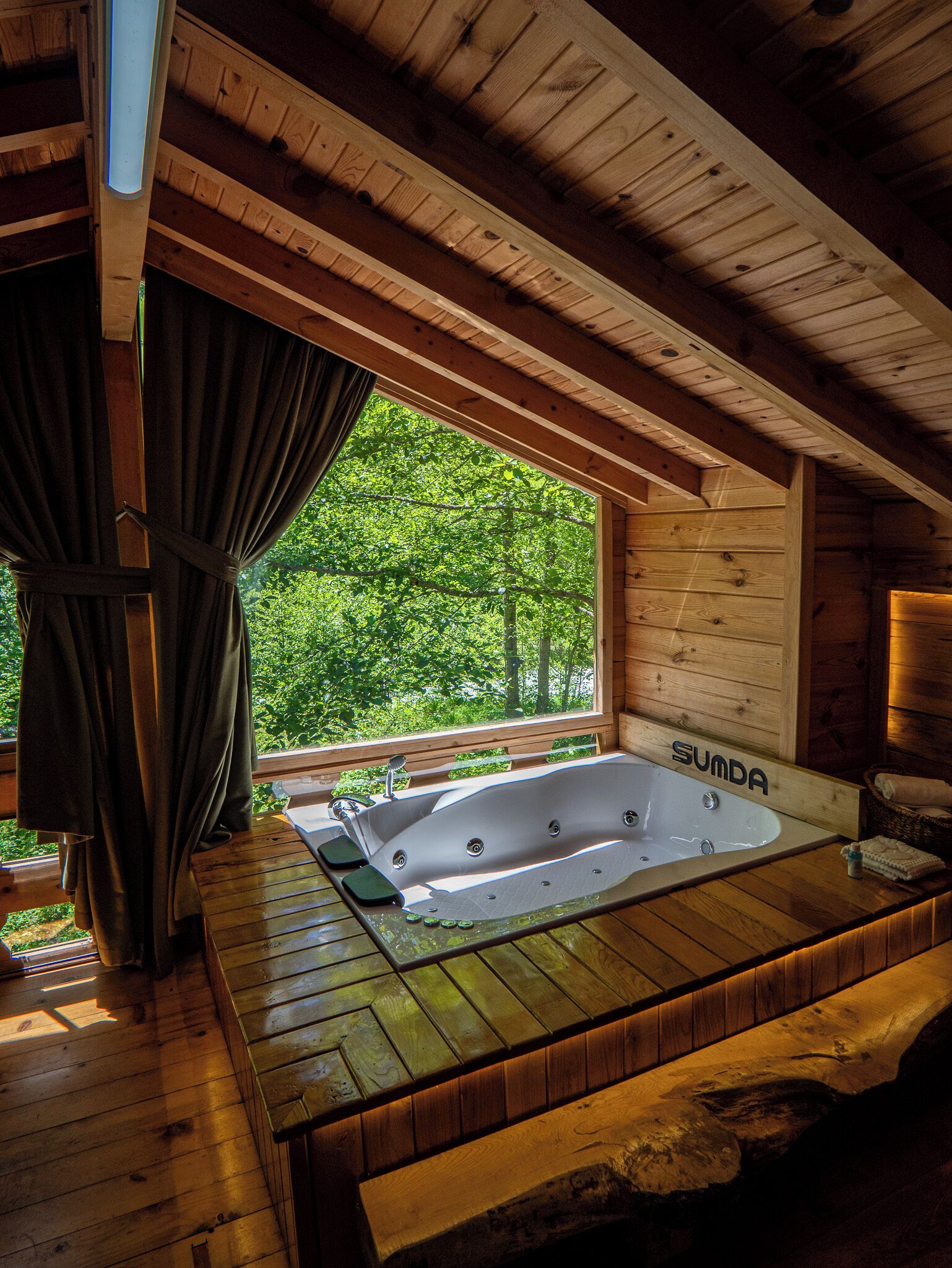 A wooden cabin with a wooden deck, a large window with curtains, and a hot tub labeled "SUMDA".