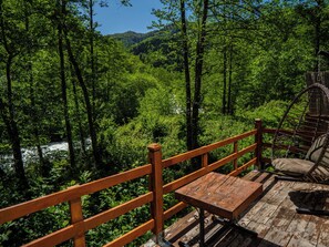 Une terrasse en bois avec un banc et une chaise en osier, offrant une vue sur une forêt verdoyante et une rivière.