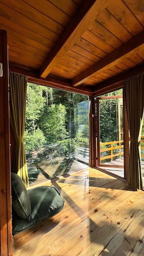 Una cabaña de madera con un ventanal grande, una terraza de madera y una zona de estar acolchada de color verde.