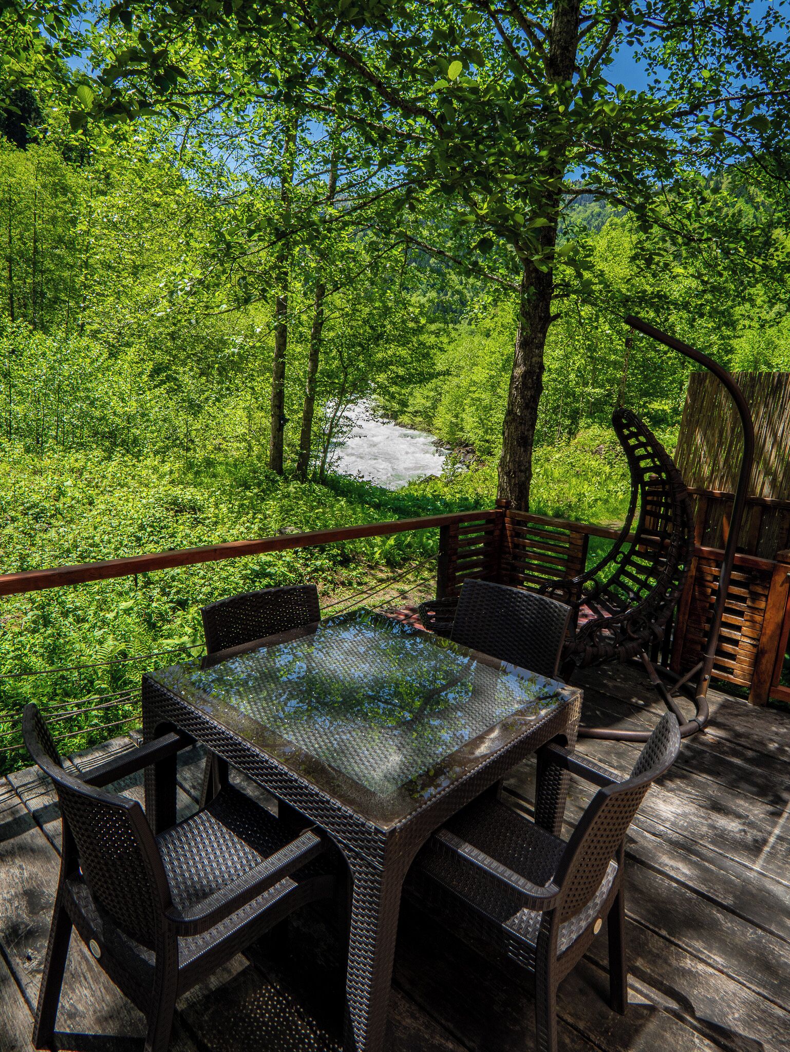 Een terras buiten met een tafel en stoelen met een glazen blad, omgeven door weelderige begroeiing en uitzicht op een rivier.