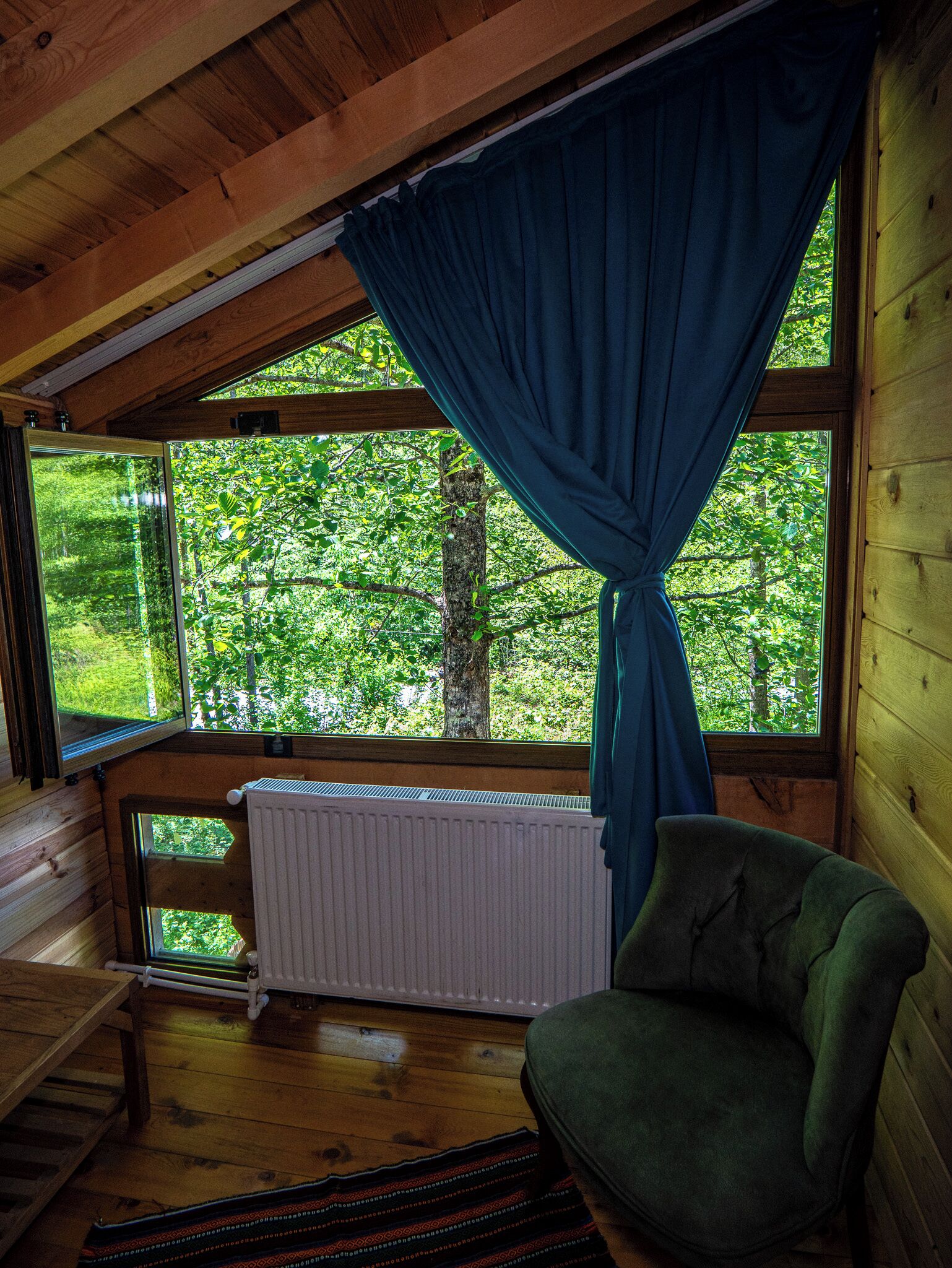 Una baita con interni in legno, vista su una foresta attraverso un'ampia finestra e una poltrona verde.