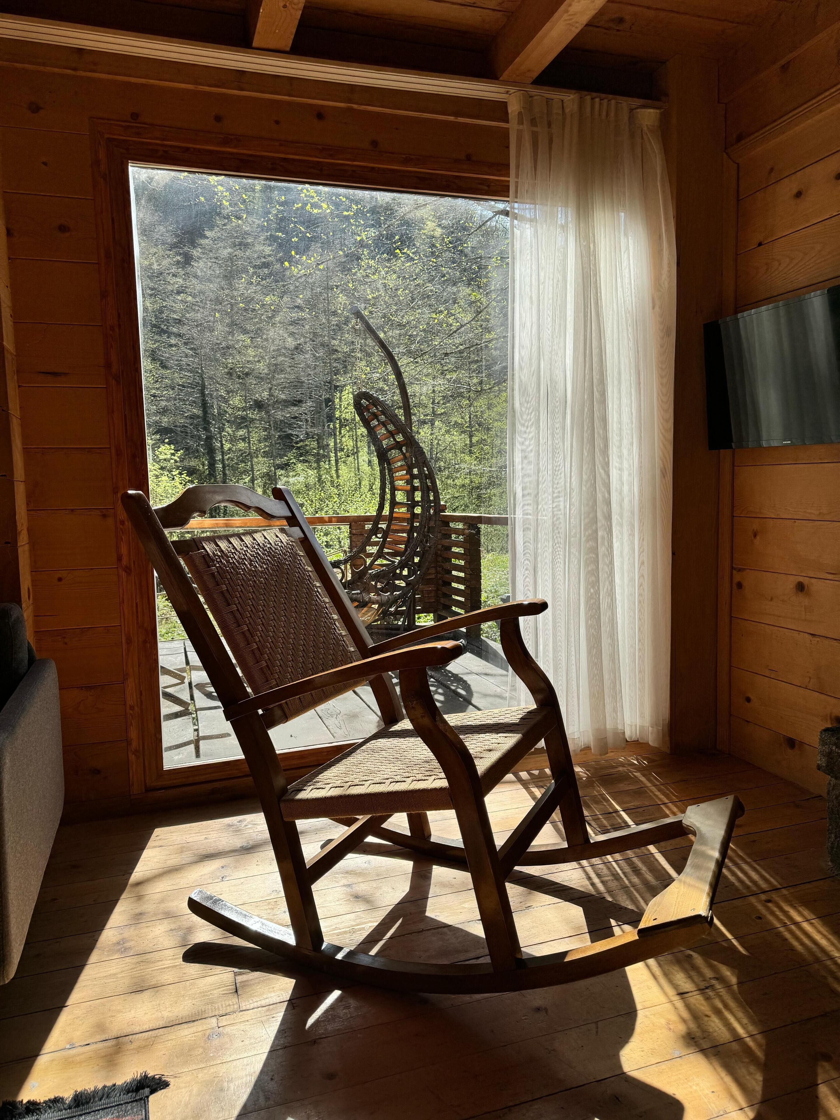 A wooden rocking chair in a cabin with a large window overlooking a forest.
