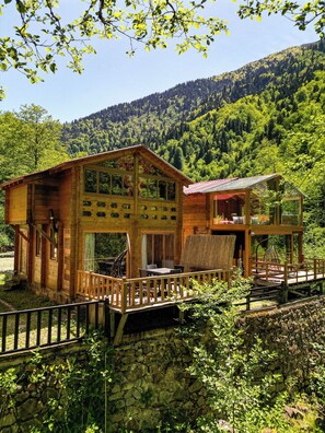 Une cabane en bois dotée d’un balcon et d’un mur en pierre, entourée d’une végétation luxuriante et surplombée par une montagne boisée.