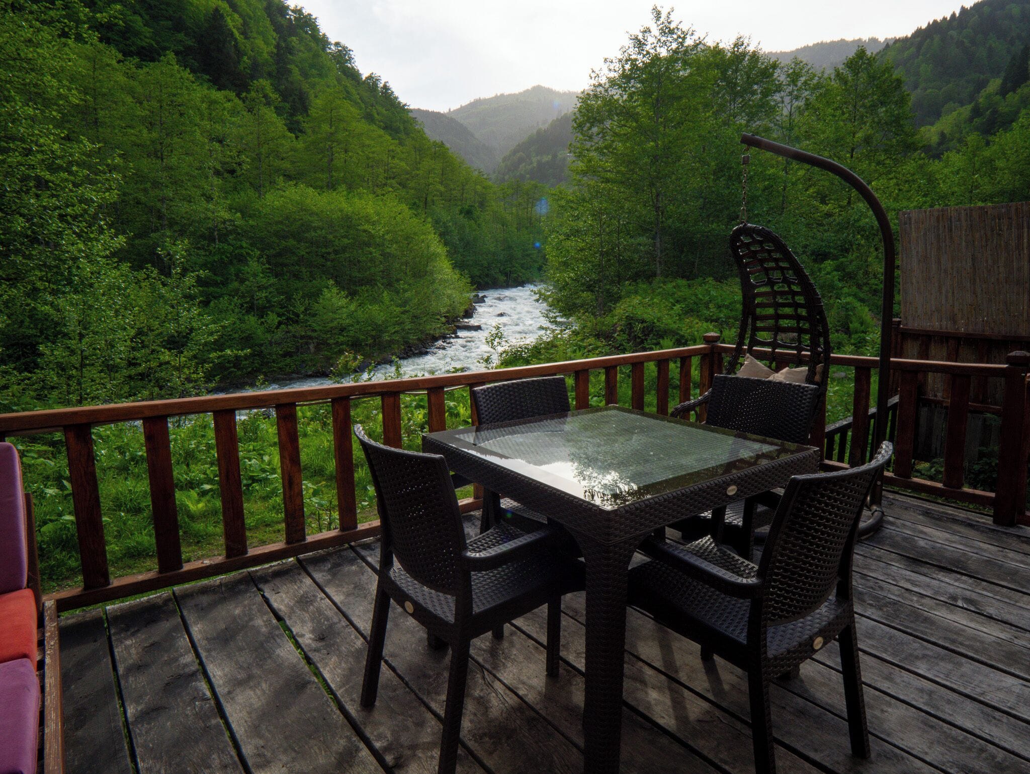 Eine Holzterrasse mit Tischplatte aus Glas und Korbstühlen, mit Blick auf einen Fluss und bewaldete Hügel.