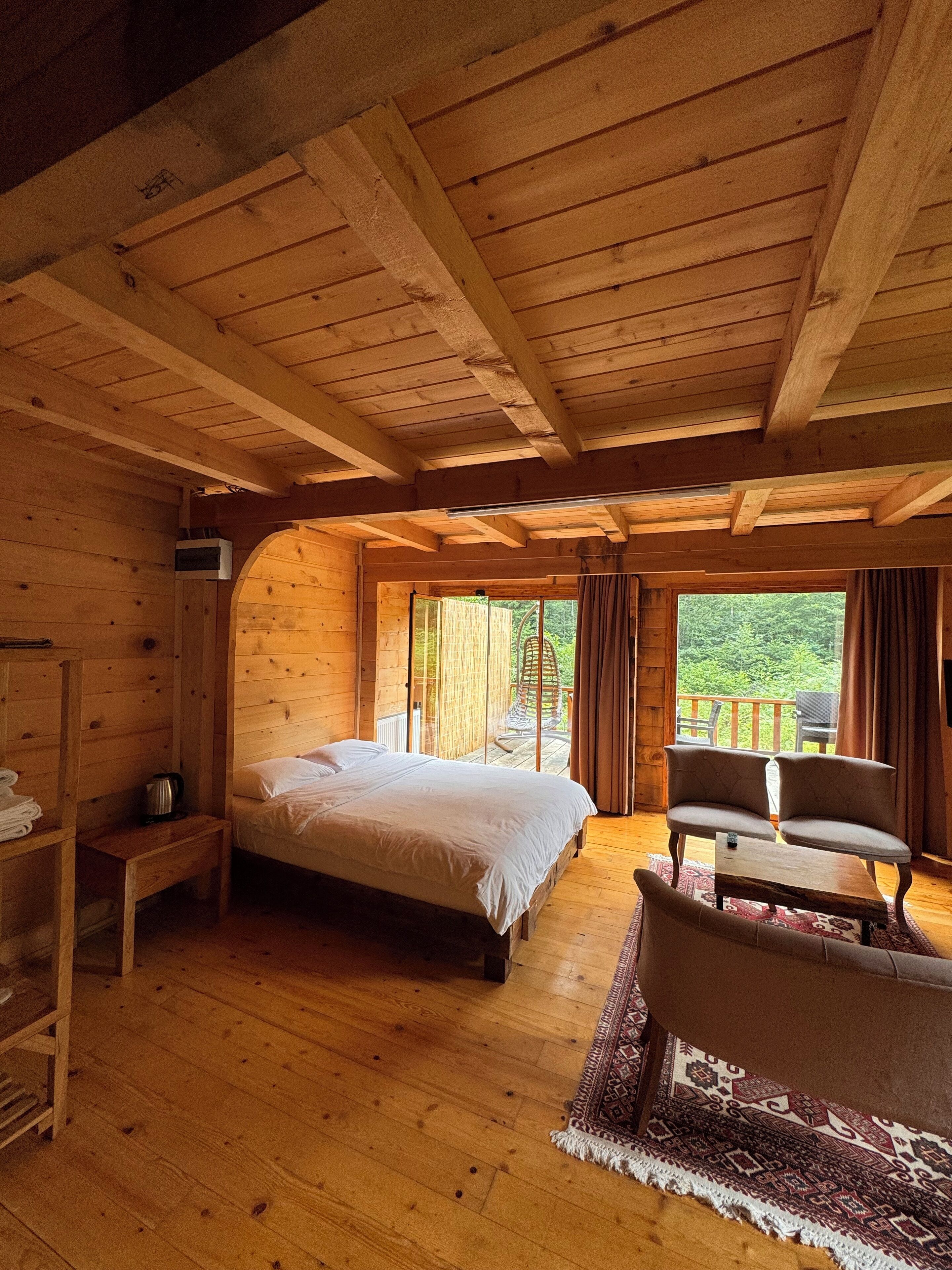 A wooden cabin bedroom with a bed, a seating area with two chairs, a small table, and a large window offering a view of the outdoors.