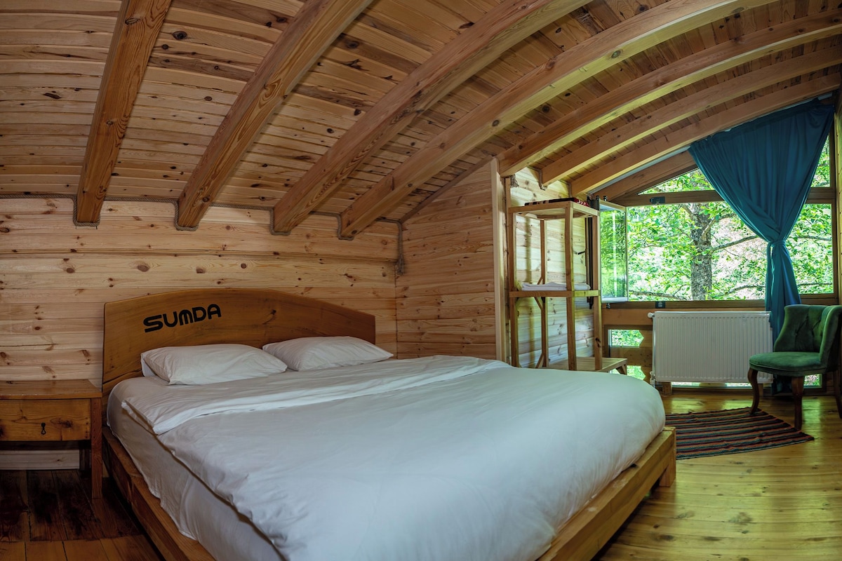A wooden cabin bedroom with a bed labeled 'Sunda', a window with curtains, and a view of greenery outside.