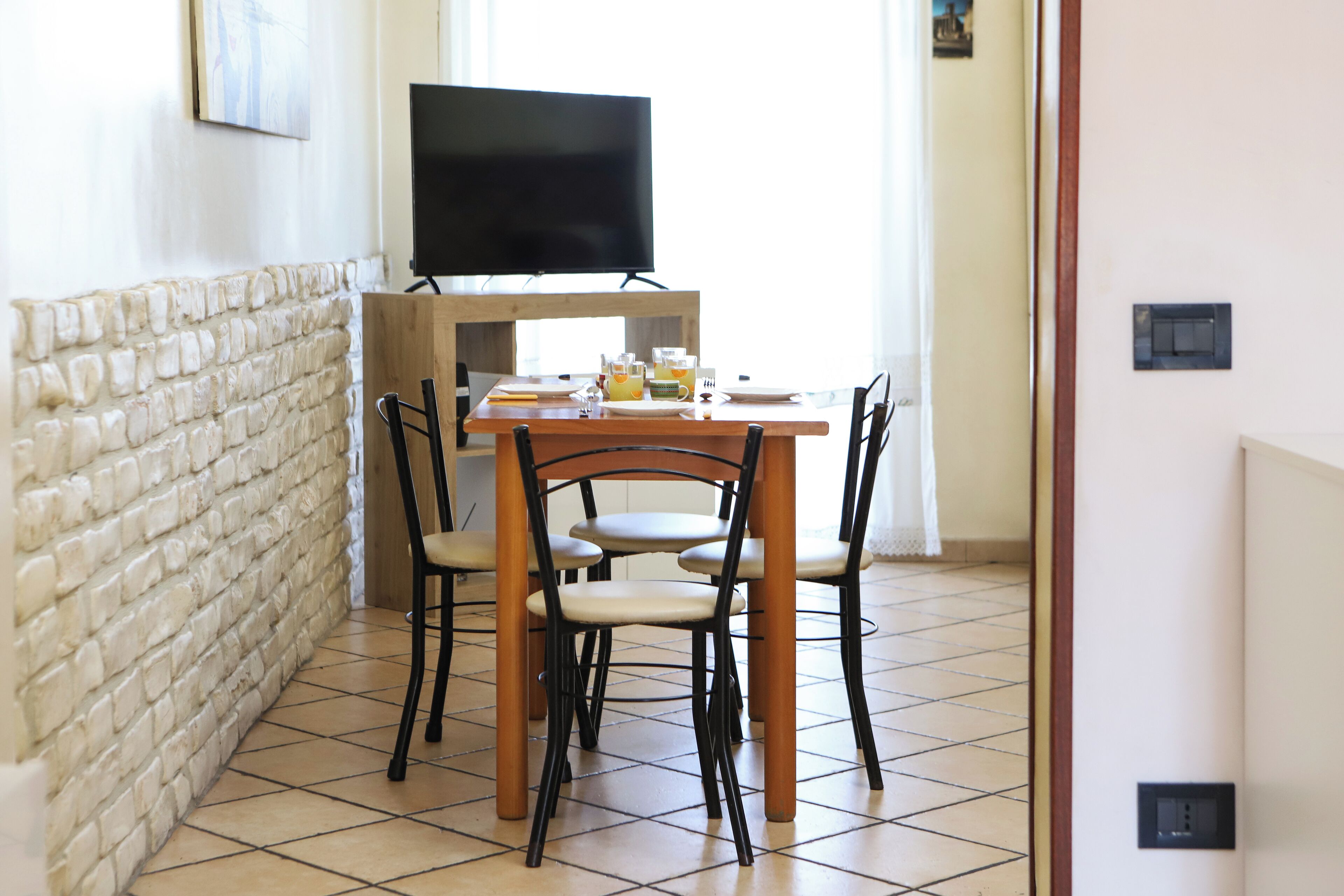 Een eetgedeelte met een houten tafel, vier stoelen, een televisie op een standaard en een bakstenen muur.