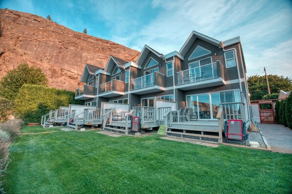 A modern multi-story house with a balcony and a deck, surrounded by a well-maintained lawn and a rocky hillside.