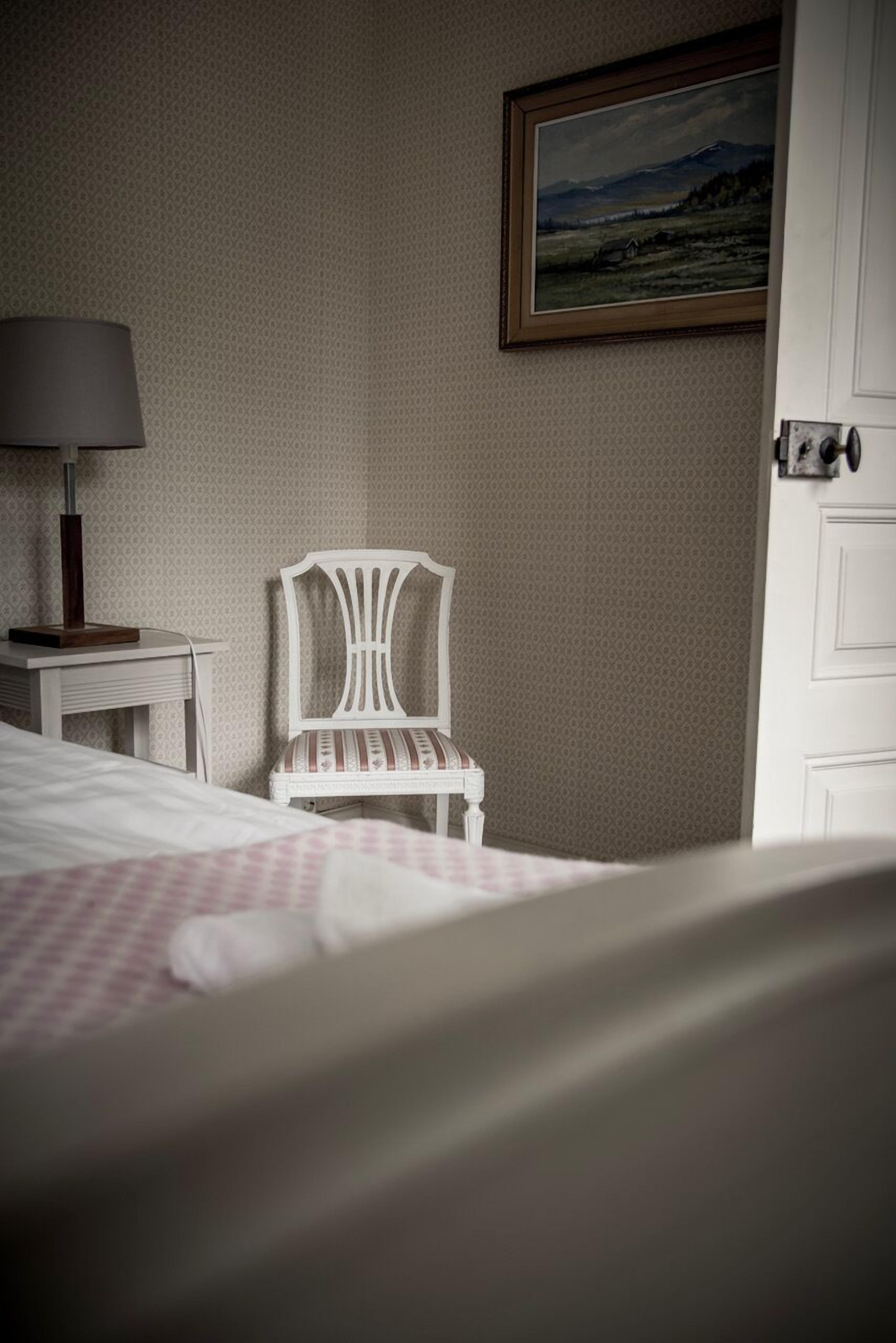 A neatly arranged bedroom with a bed, a chair, a lamp, and a painting on the wall.