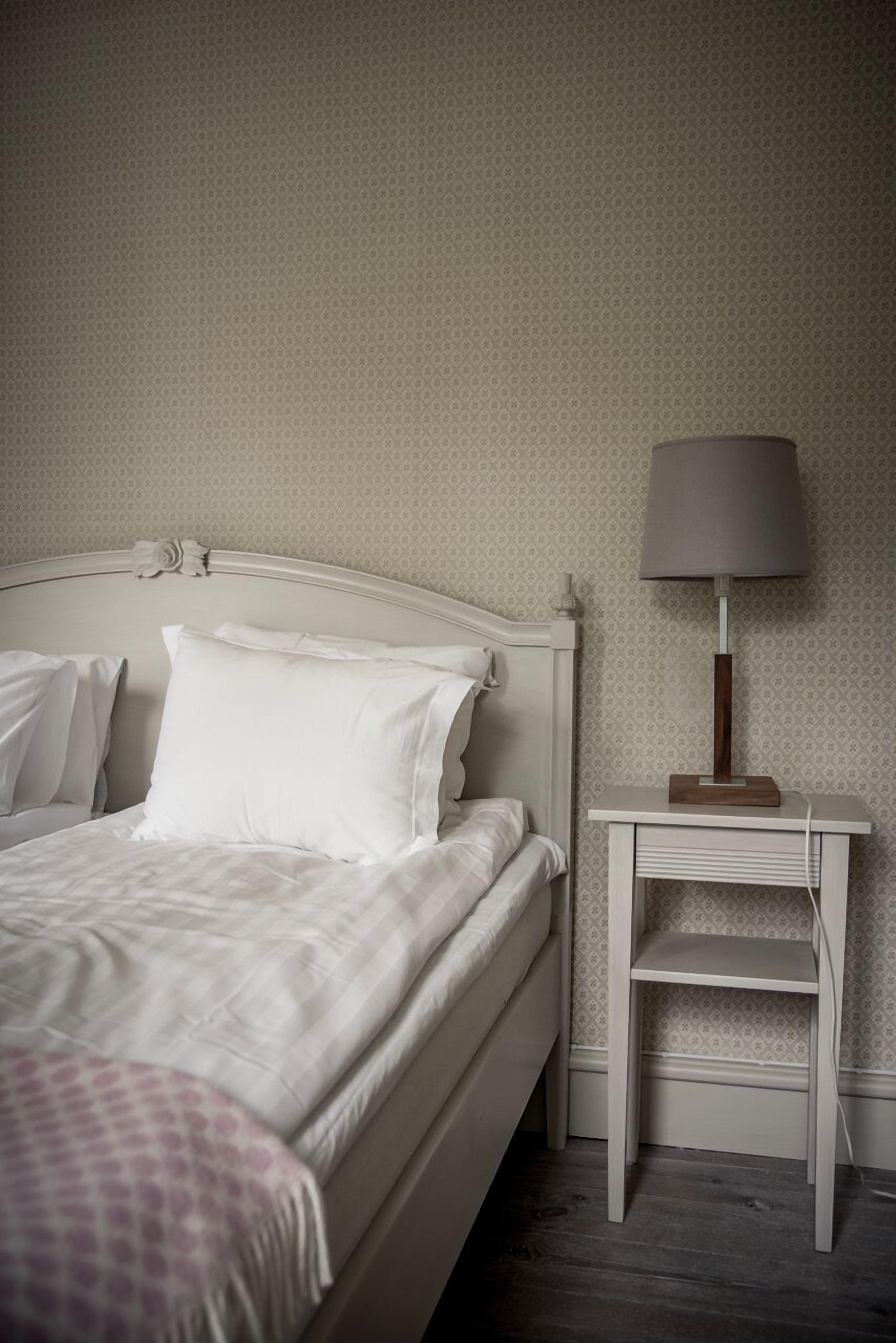 A neatly made bed with white linens, a bedside table with a lamp, and a patterned wall.