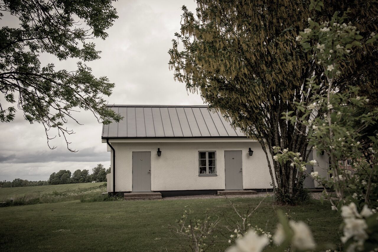 A white building with a grey roof, two doors, and a window, surrounded by grass and trees.