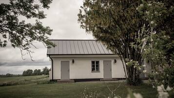 A white building with a grey roof, two doors, and a window, surrounded by grass and trees.
