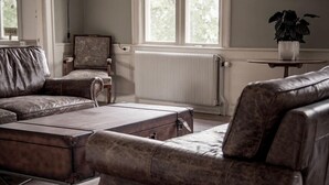 A room with a large window, a radiator, a leather sofa, a small table with a plant, and a framed mirror on the wall. - Ruders Egendom (Tibro)