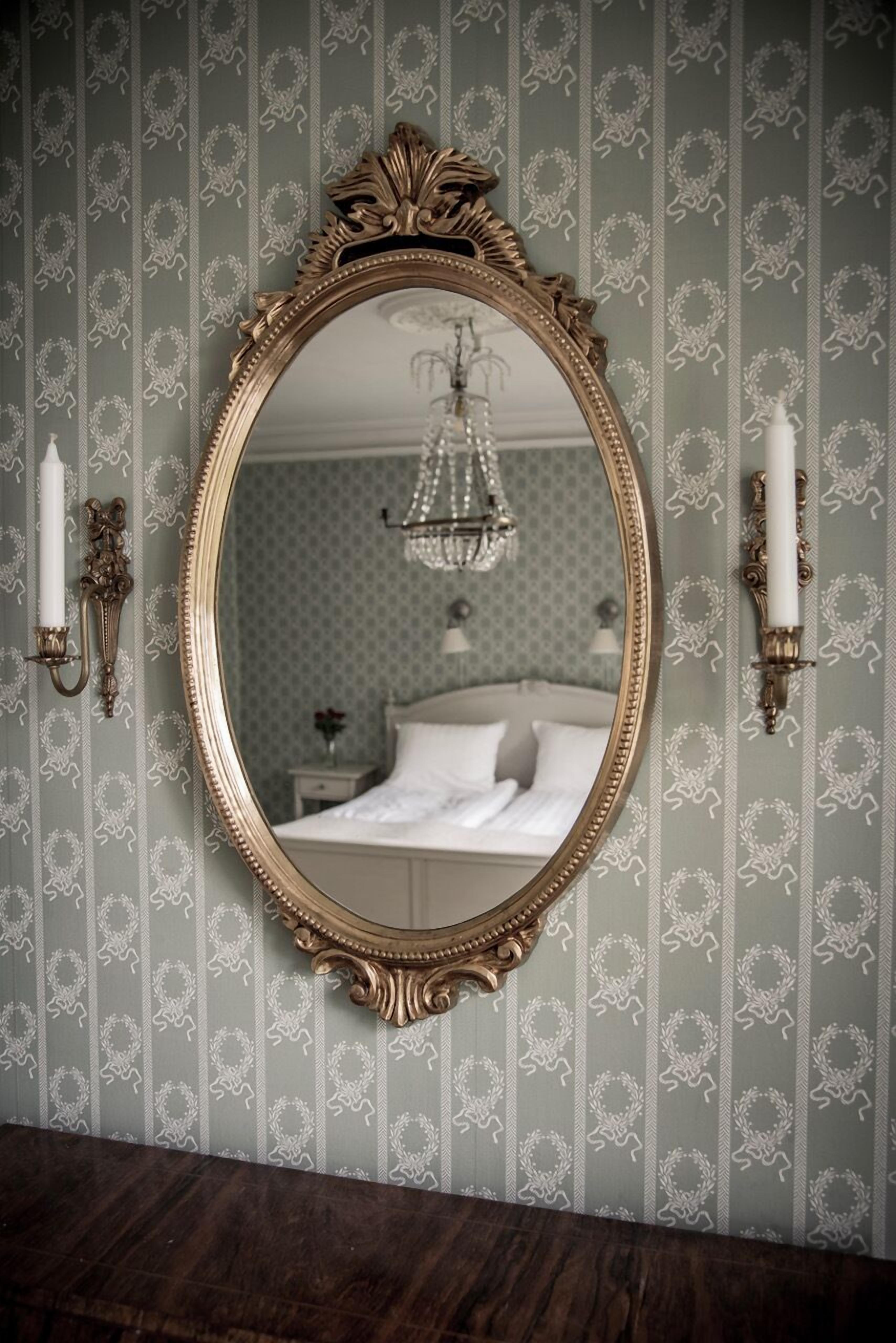 A mirror with an ornate gold frame, set against a wallpapered wall with a repeating pattern.