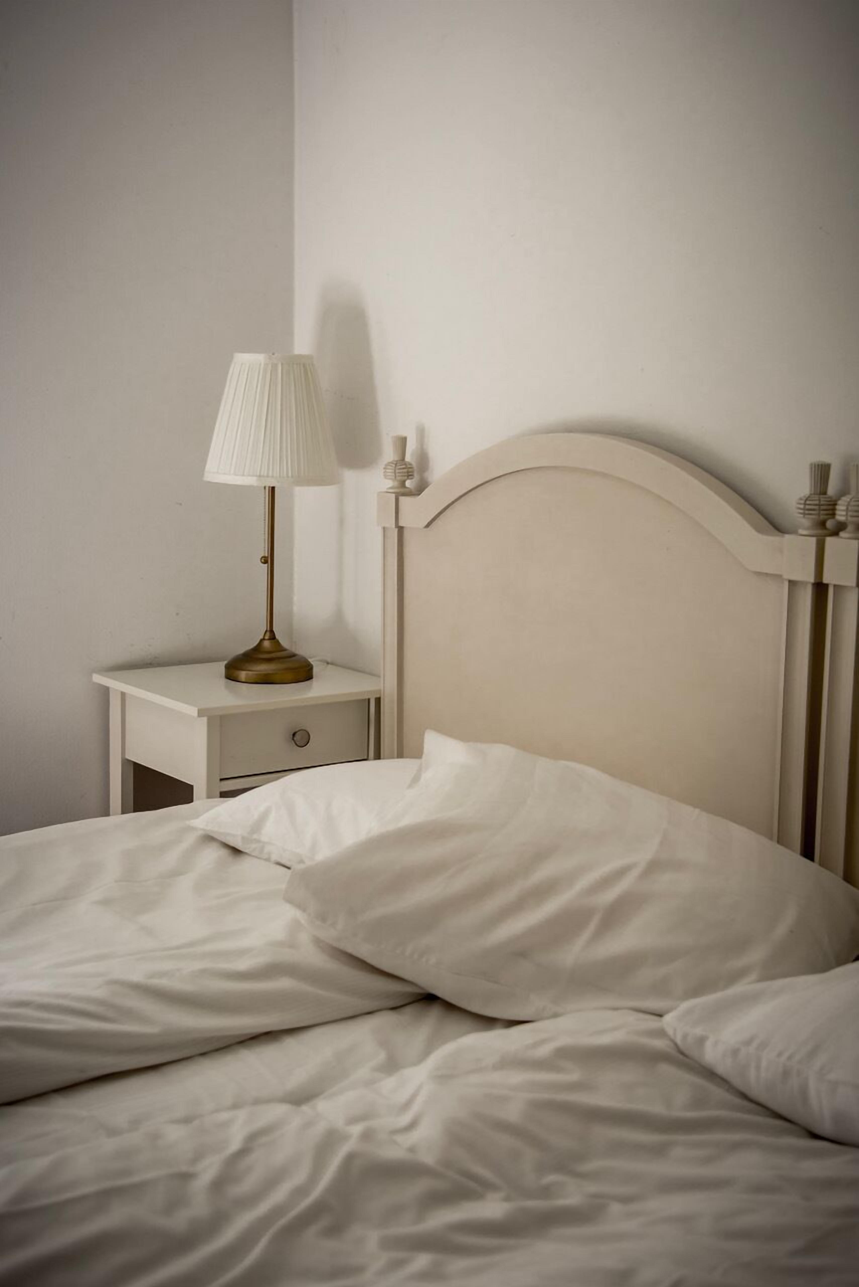 En pænt redt seng med hvide sengetæpper og et enkelt natbord med en lampe.