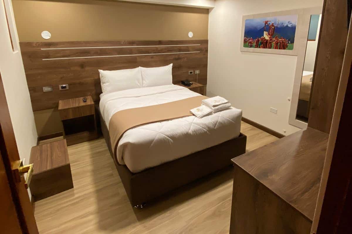 A hotel room with a bed, bedside tables, a nightstand, a wooden headboard, a ceiling light, and a painting on the wall.