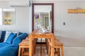 Una sala de estar moderna con un sofá azul, una mesa de comedor de madera dispuesta con cuencos blancos y copas de vino, y un espejo que refleja la habitación.