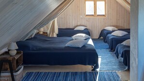 Una habitación acogedora con paneles de madera, con dos camas, cada una con colcha azul y almohadas blancas. Una ventana deja entrar luz natural, y hay un tapete azul a rayas en el suelo.