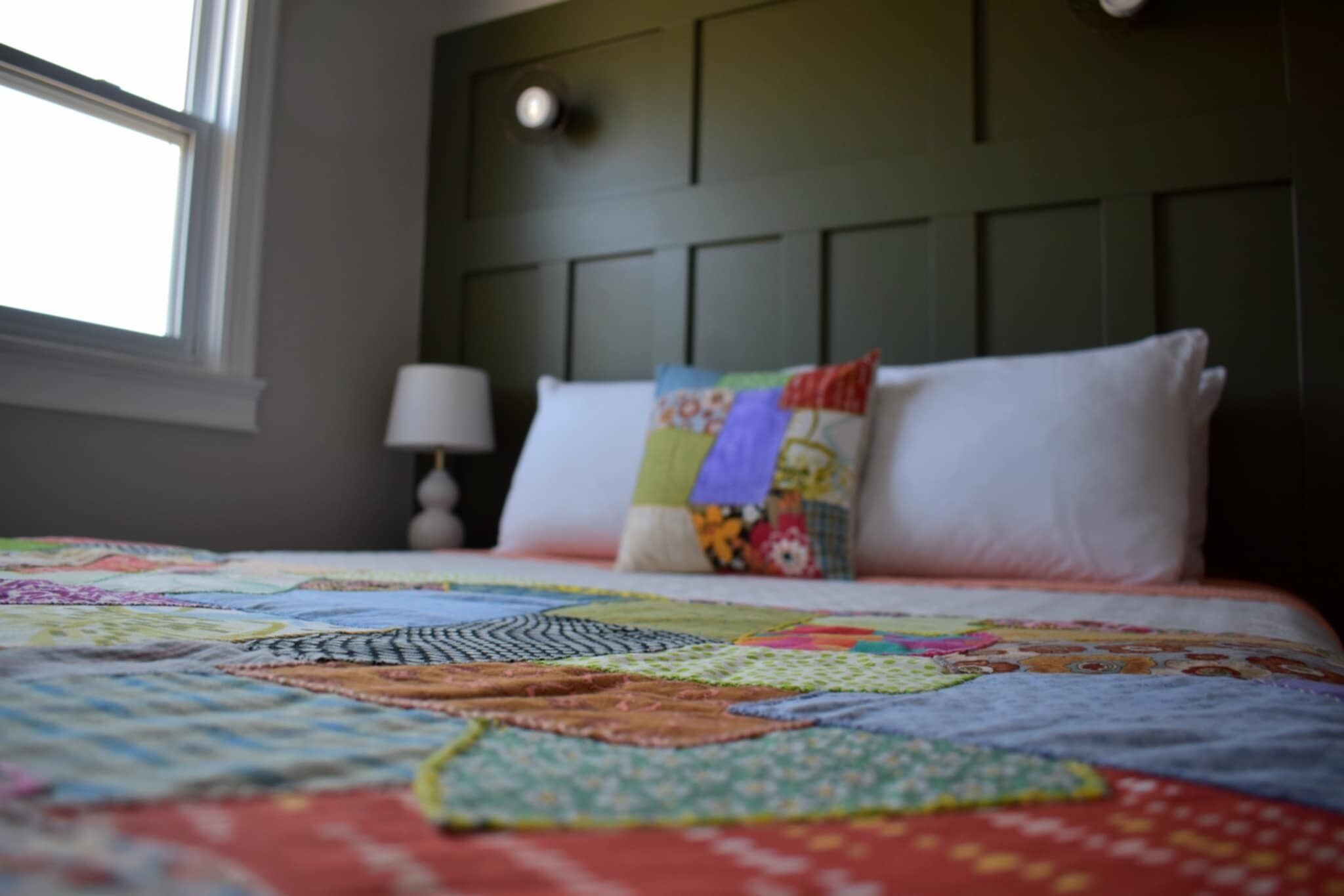 A bed with a colorful quilt, a window, and a wall-mounted lamp.