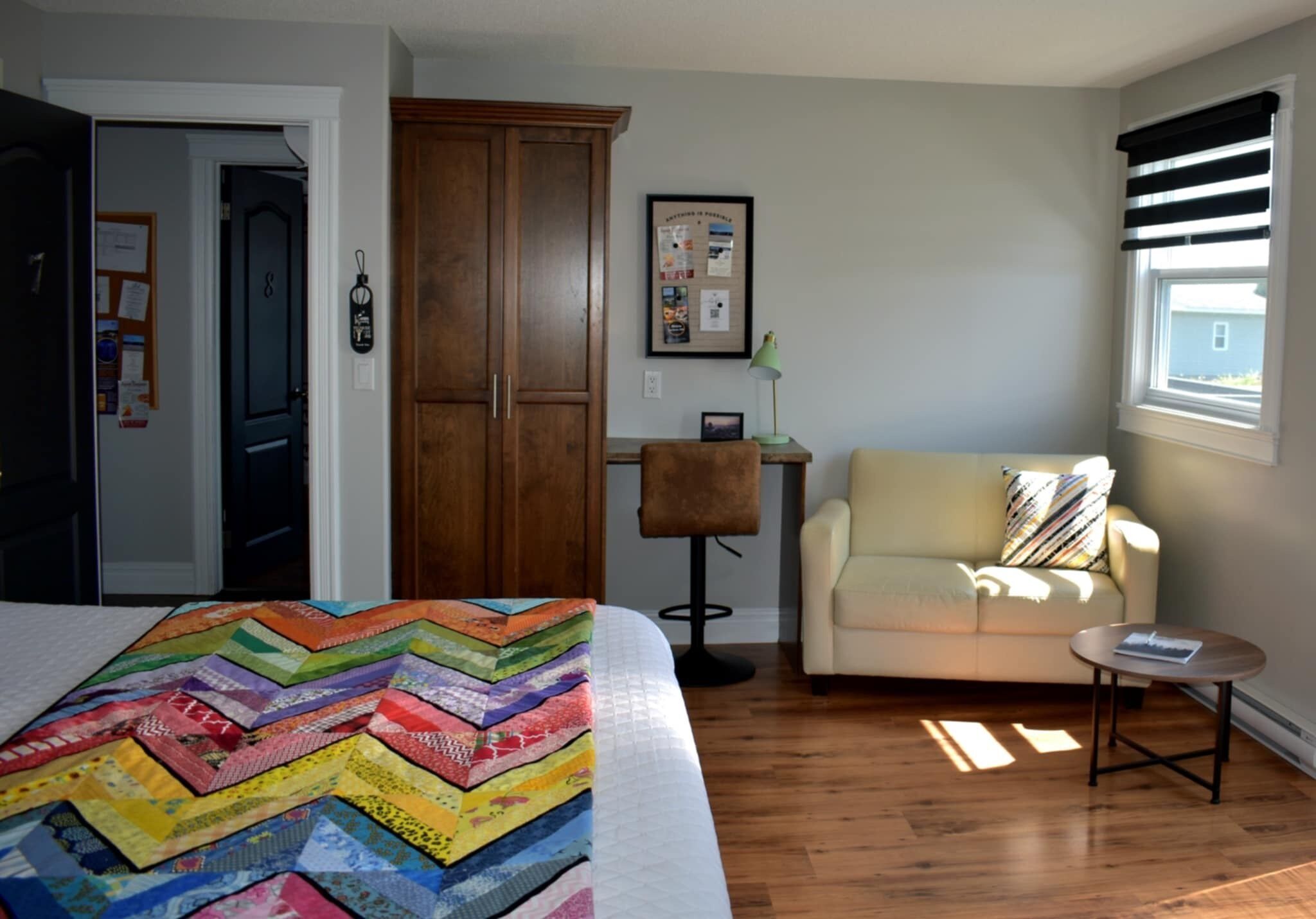 Un dormitorio con cama, un armario de madera, una mesita de noche con lámpara, un sofá y una ventana con persianas.