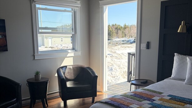 Une chambre avec un lit, une chaise, une petite table, un ventilateur de plafond et une vue sur l’extérieur, visible à travers la porte.