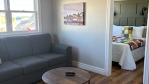 Un salon moderne avec un canapé gris, une table basse ronde en bois et un lit recouvert d’une courtepointe aux couleurs vives.