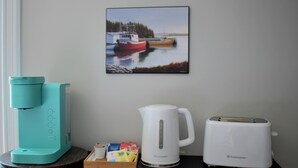 Un coin cuisine avec une cafetière turquoise, une bouilloire électrique blanche et un grille-pain blanc posés sur un plan de travail foncé. On peut également apercevoir un petit plateau en bois contenant des sachets de thé et une tasse.